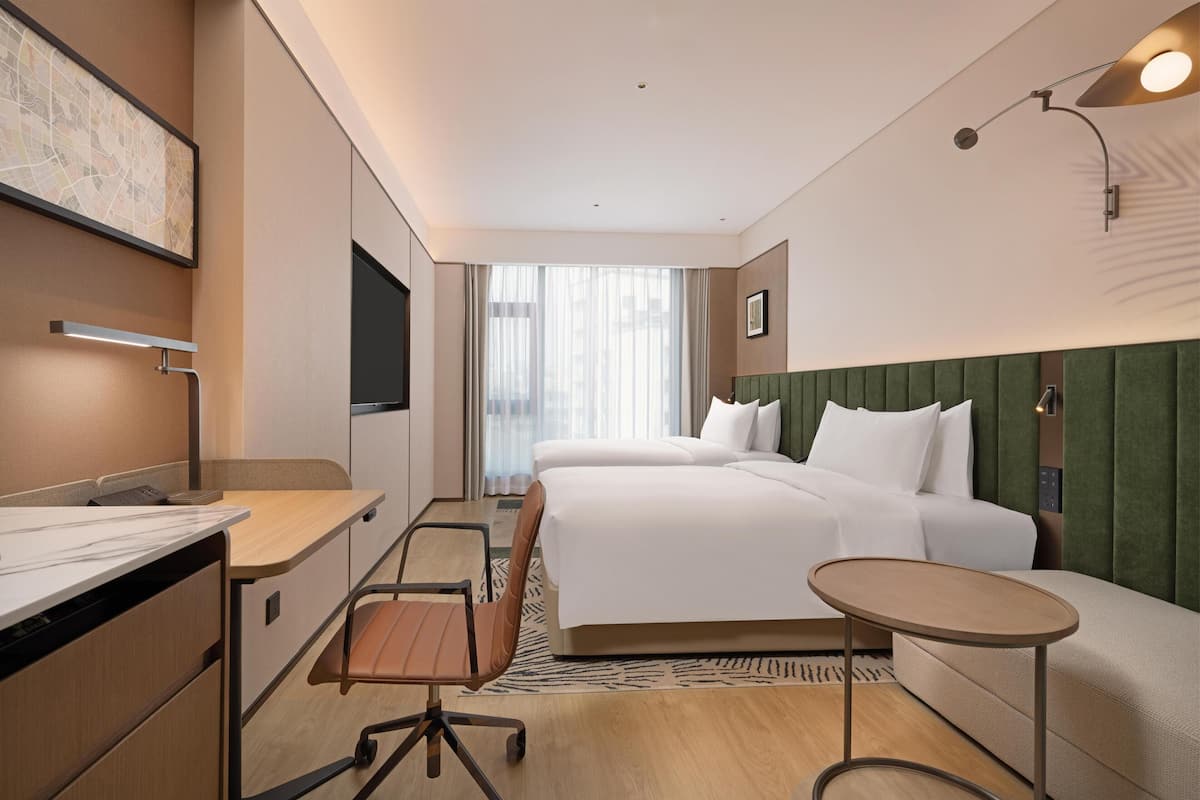Habitación de hotel con escritorio, dos camas, una silla, una mesita, televisor y ventana con cortinas.
