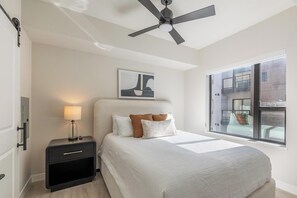 Une chambre à coucher comprenant un lit, une table de chevet, une lampe, un ventilateur de plafond, une fenêtre et un tableau accroché au mur.