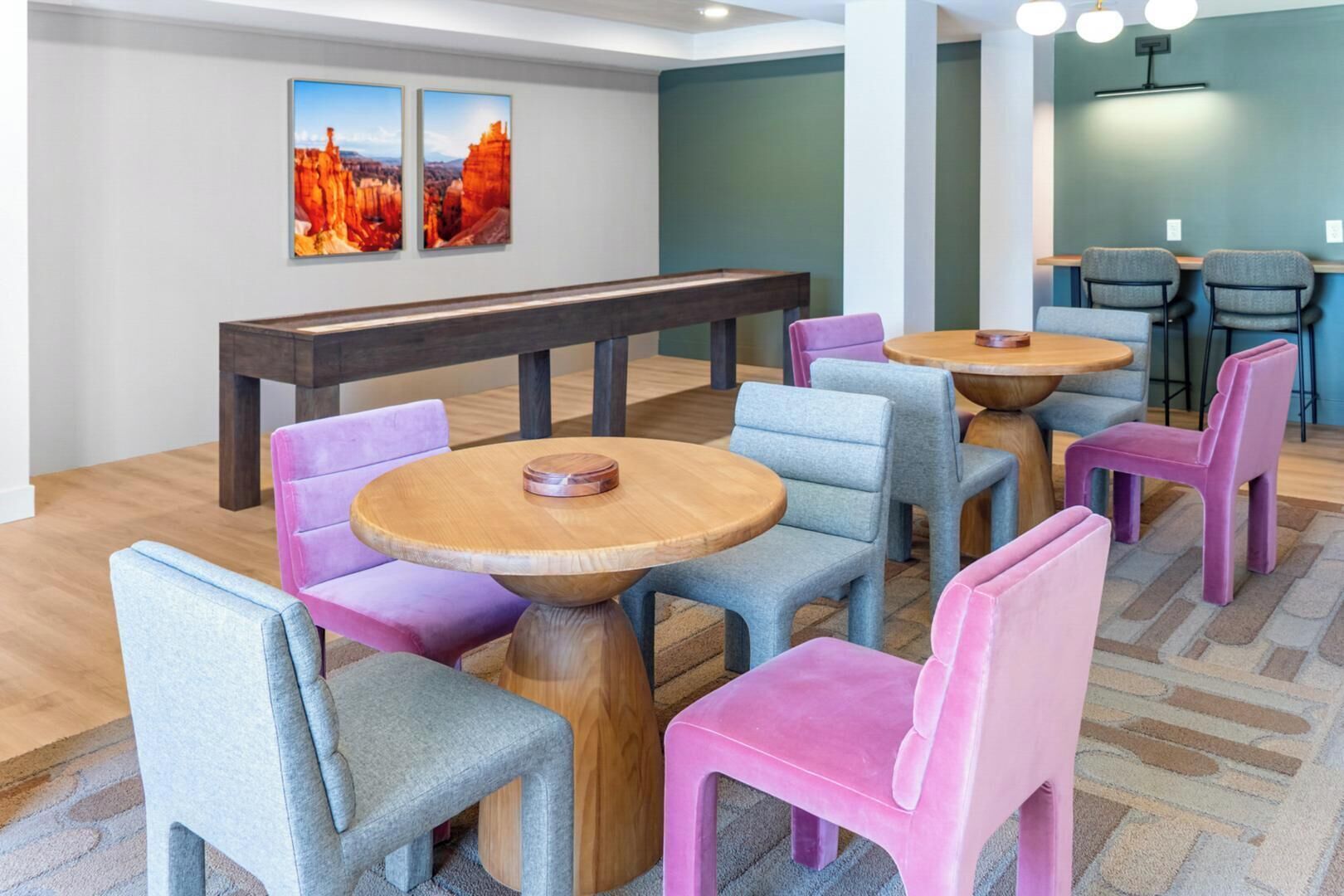 Um lobby de hotel com mesas redondas de madeira e cadeiras coloridas, um banco e duas pinturas penduradas na parede.