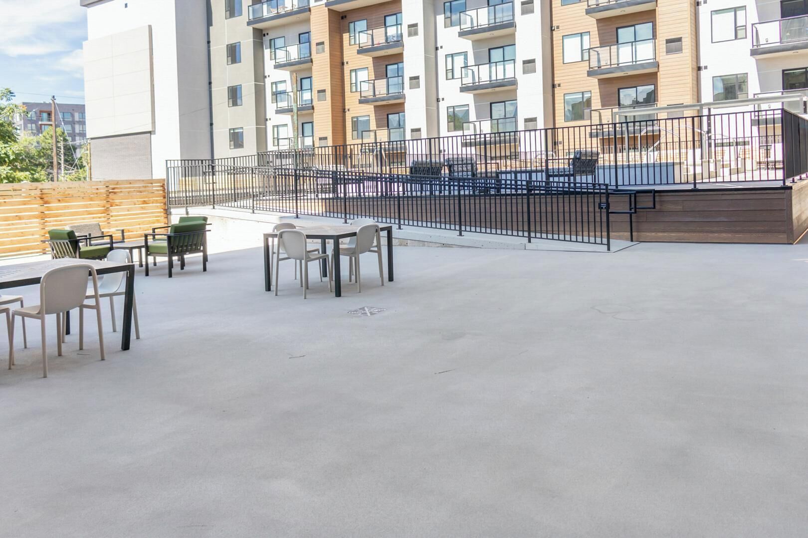 Een terras buiten met tafels en stoelen, omgeven door een metalen hek, en een modern gebouw op de achtergrond.