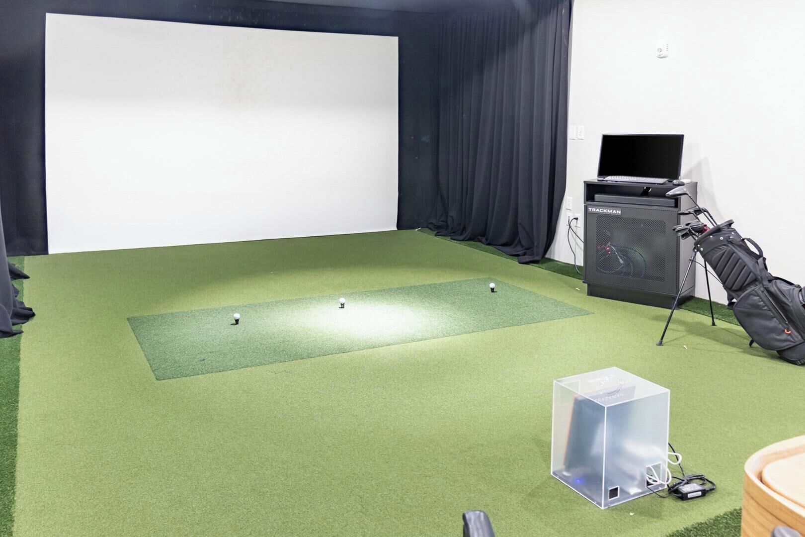 A room with a green carpet, a projector screen, a golf bag, and a speaker.