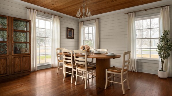 A dining room with a wooden table and chairs, large windows, and a chandelier.