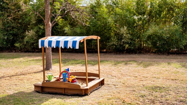 Una casita de juegos de madera con un toldo azul y blanco a rayas, ubicada en un c ésped con árboles al fondo.