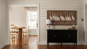 Un coin repas avec une table et des chaises en bois, une console noire surmontée d’une lampe, et une œuvre d’art représentant des oiseaux, accrochée au mur.