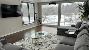 Ein modernes Wohnzimmer mit einem großen, grauen Ecksofa, einem Glastisch und Blick auf eine verschneite Landschaft durch große Fenster.