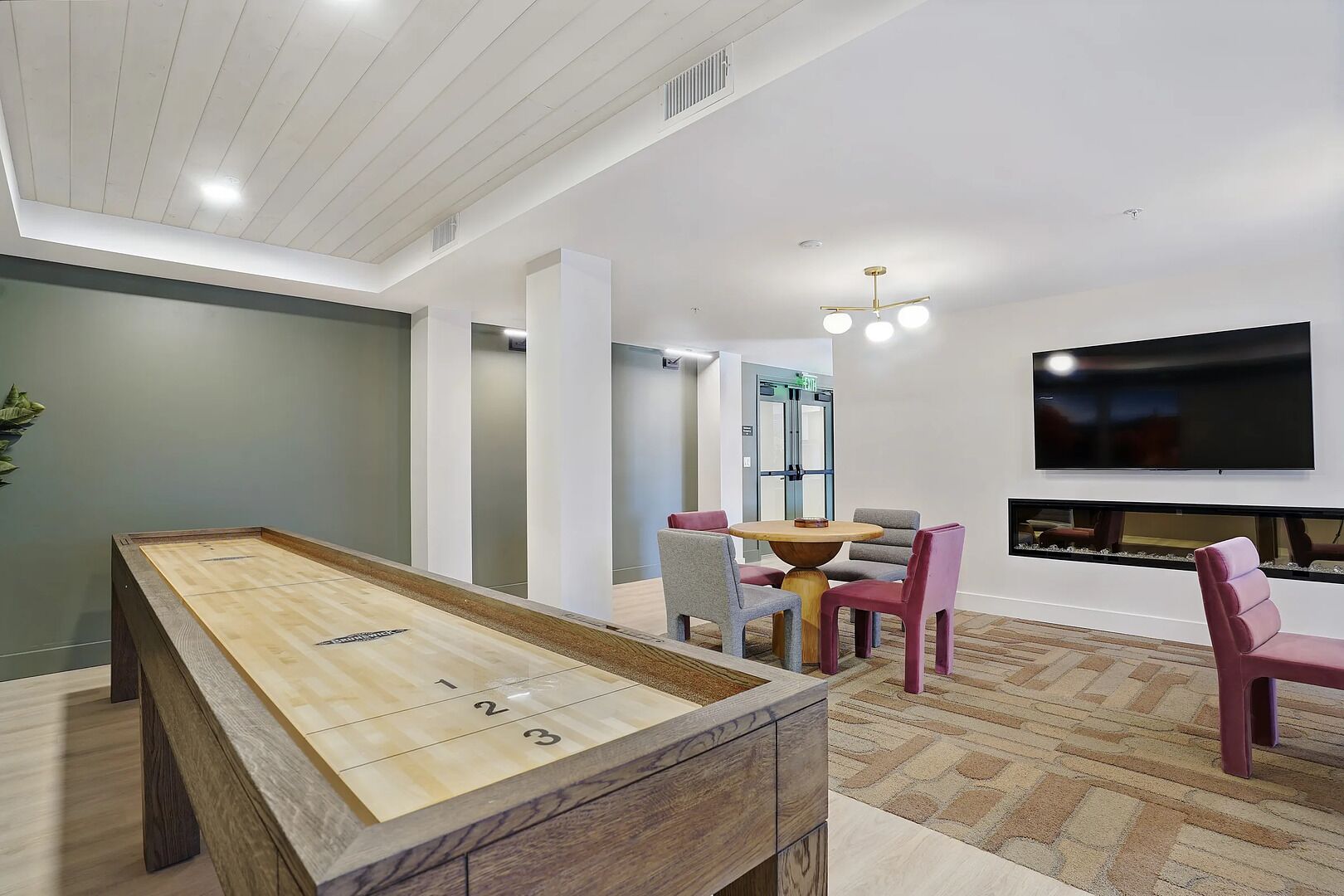 A modern living room with a shuffleboard table, a dining area with chairs, a fireplace, and a flat-screen TV mounted on the wall.