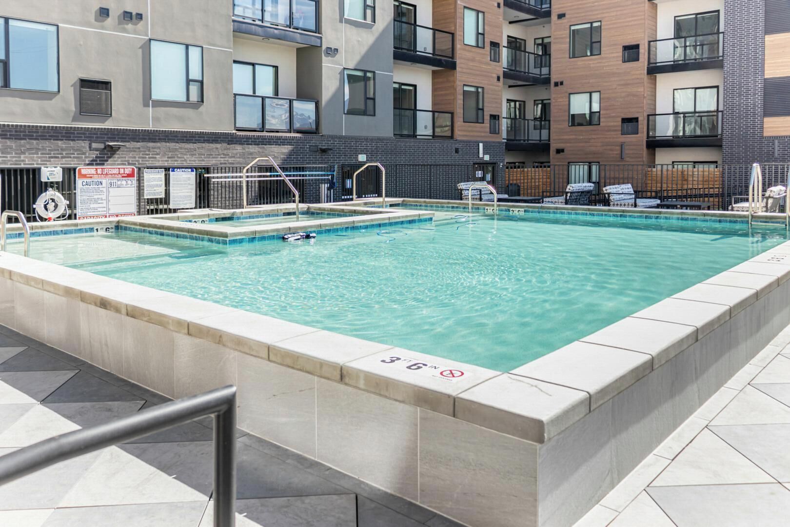 A rectangular swimming pool with a tiled edge, surrounded by a metal fence, and located in a multi-story residential building with balconies.