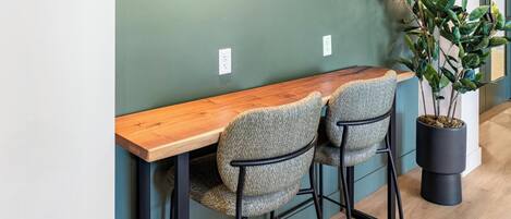 A wooden bar counter with two upholstered chairs, a potted plant, and a green wall with a power outlet.