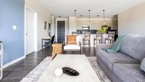 Un salon moderne avec un canapé gris, une table basse et un coin repas comprenant des chaises, et une cuisine en arrière-plan.