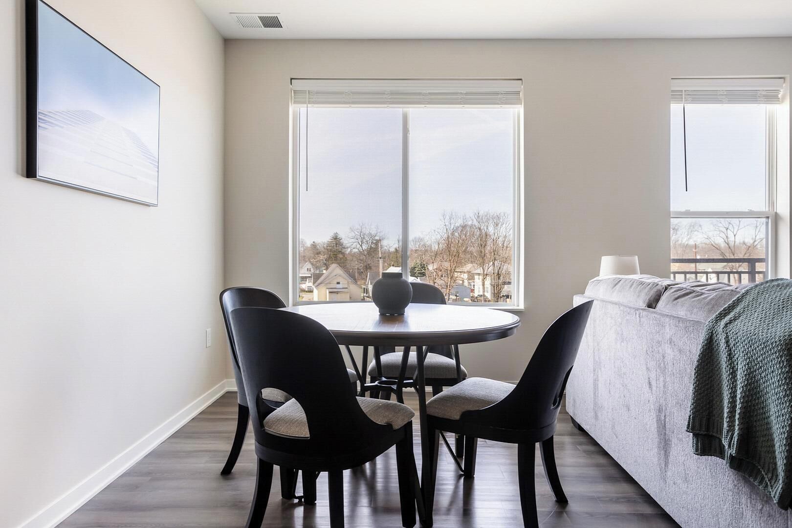 A room with a round table, four chairs, a sofa, and two large windows offering a view of houses.