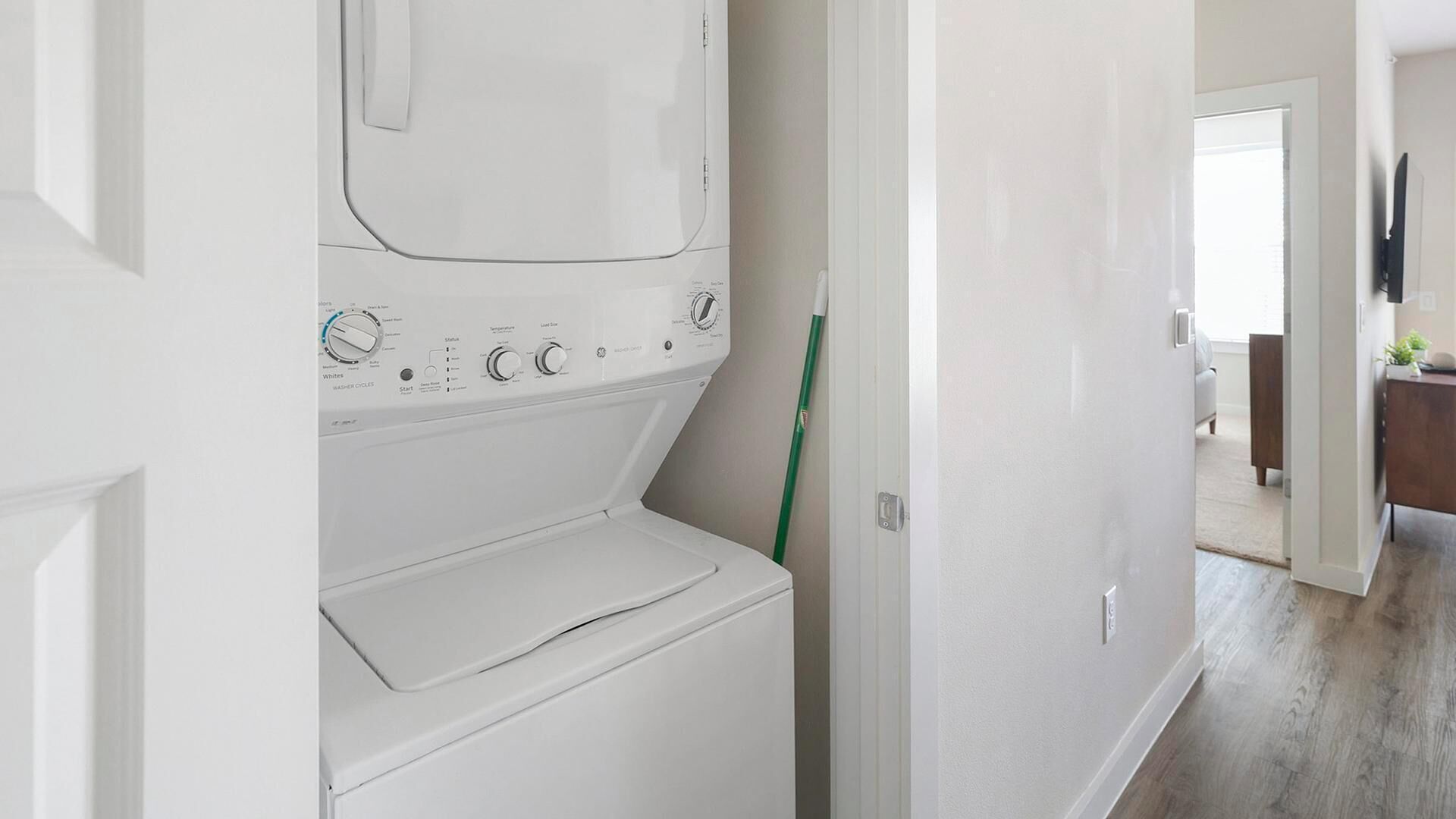 Un lavadero con lavadora y secadora de carga frontal, una escoba apoyada en la pared, y un acceso a una sala de estar con sofá y televisor.