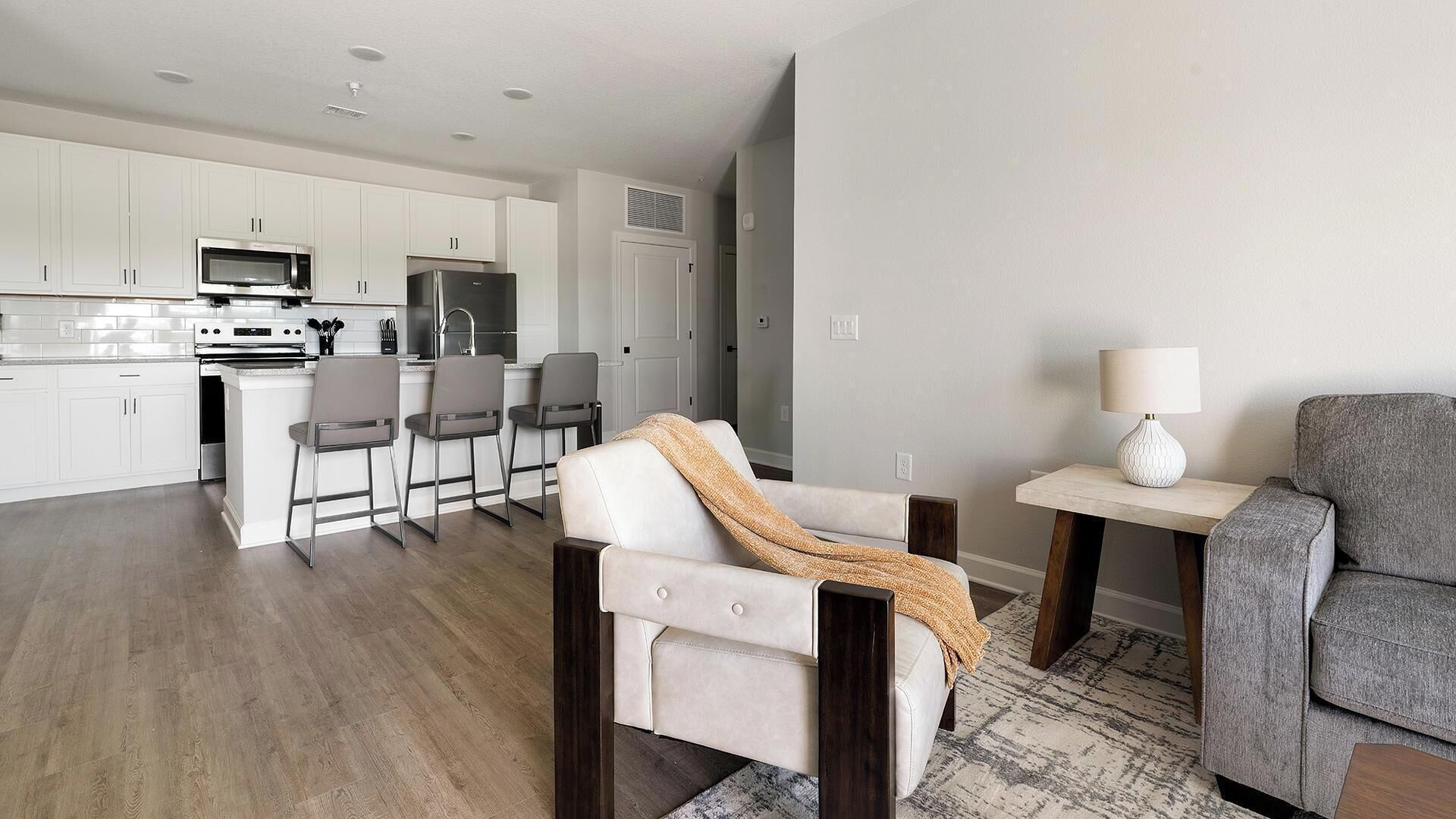 Une cuisine moderne avec des meubles blancs et des appareils en acier inoxydable, un coin repas avec un canapé blanc et un fauteuil gris, ainsi qu’une petite table avec une lampe.