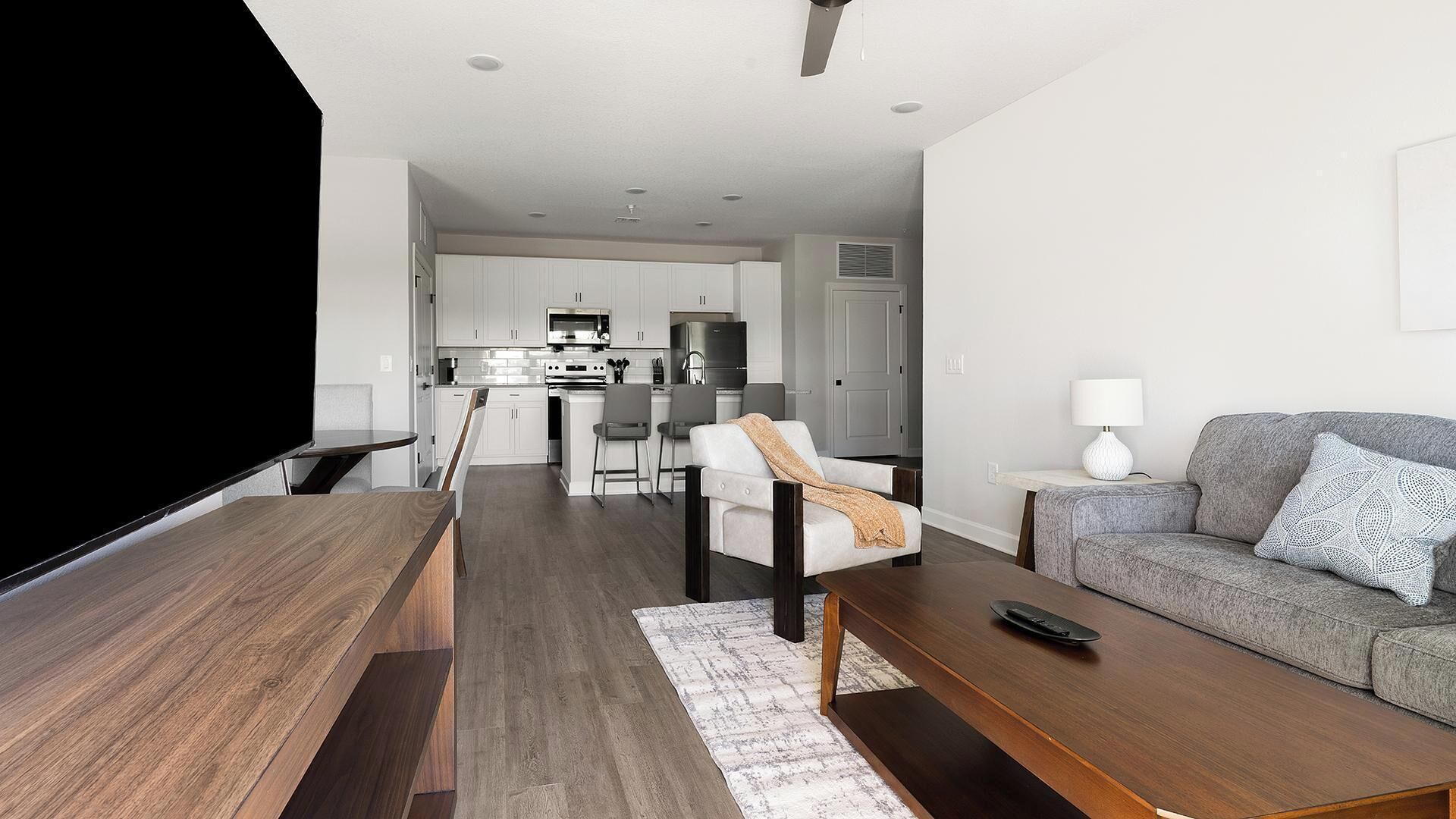A modern living room with a flat-screen TV, a grey sofa, a wooden coffee table, and a dining area with a kitchen in the background.