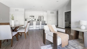 Eine moderne Küche mit weißen Schränken, einer zentralen Kochinsel und Edelstahlgeräten. Es gibt einen Essbereich mit einem Holztisch und Stühlen, sowie eine Wohnarea mit einem Sofa und einem Couchtisch.