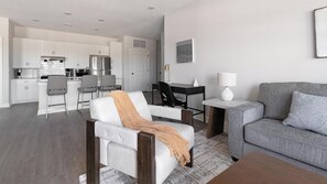 Un salon moderne avec un canapé gris, un fauteuil blanc agrémenté d’un plaid, une table basse et un coin repas avec une table et des chaises aux couleurs sombres.