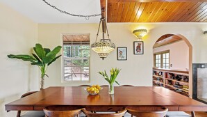 A dining area with a wooden table, grey cushioned chairs, a hanging light fixture, and a bookshelf in the background.