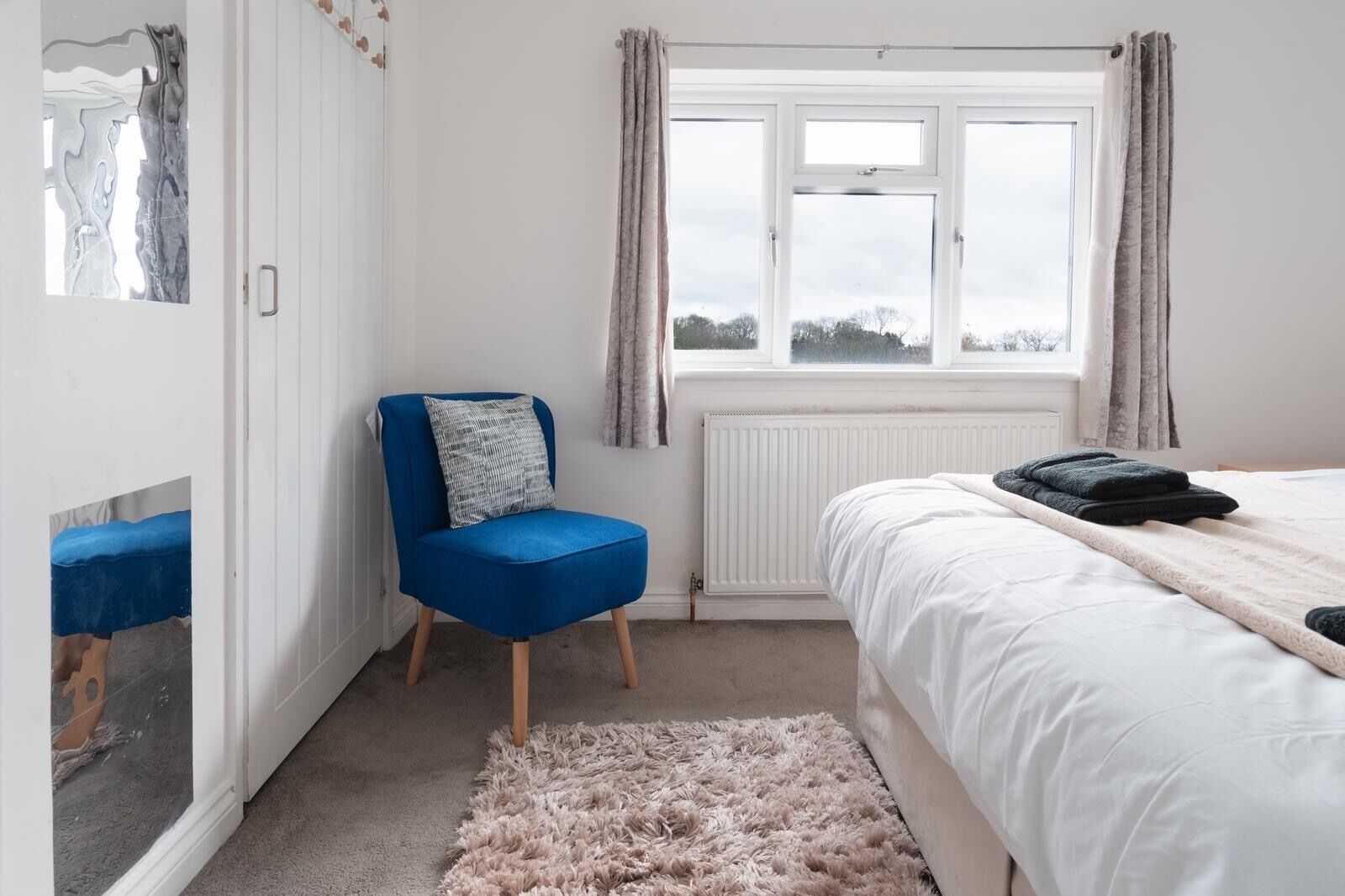 A bedroom with a bed, a blue chair, a fluffy rug, and a window with curtains.