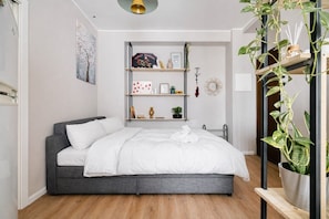 Une chambre moderne avec un lit gris, du linge de lit blanc, une étagère murale agrémentée d’objets décoratifs et une plante en pot.