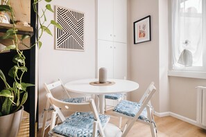 Een kamer met een ronde witte tafel, vier witte stoelen met gestreepte kussens, een potplant, een geometrisch kunstwerk aan de muur, een lijst met een foto, en een raam met dunne gordijnen.