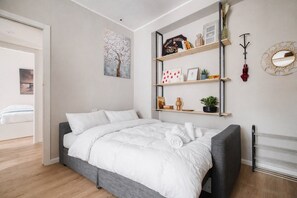 Een moderne slaapkamer met een bed, een wandplank met boeken en decoratieve objecten, en een schilderij van een boom.