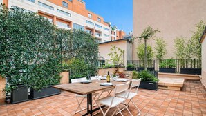 Eine Terrassensitzgruppe im Freien mit Holztisch, gedeckt für ein Essen, umgeben von Blumentöpfen und Blick auf eine Stadtlandschaft.