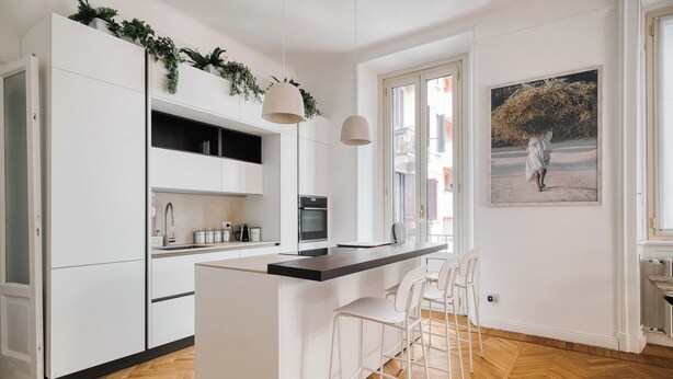 Eine moderne Küche mit weißen Schränken, einer zentralen Kochinsel mit Barhocker, einer Hängeleuchte und einem großen Fenster mit Außenblick.