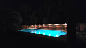 A swimming pool at night with lights around it and a wooden fence in the background.