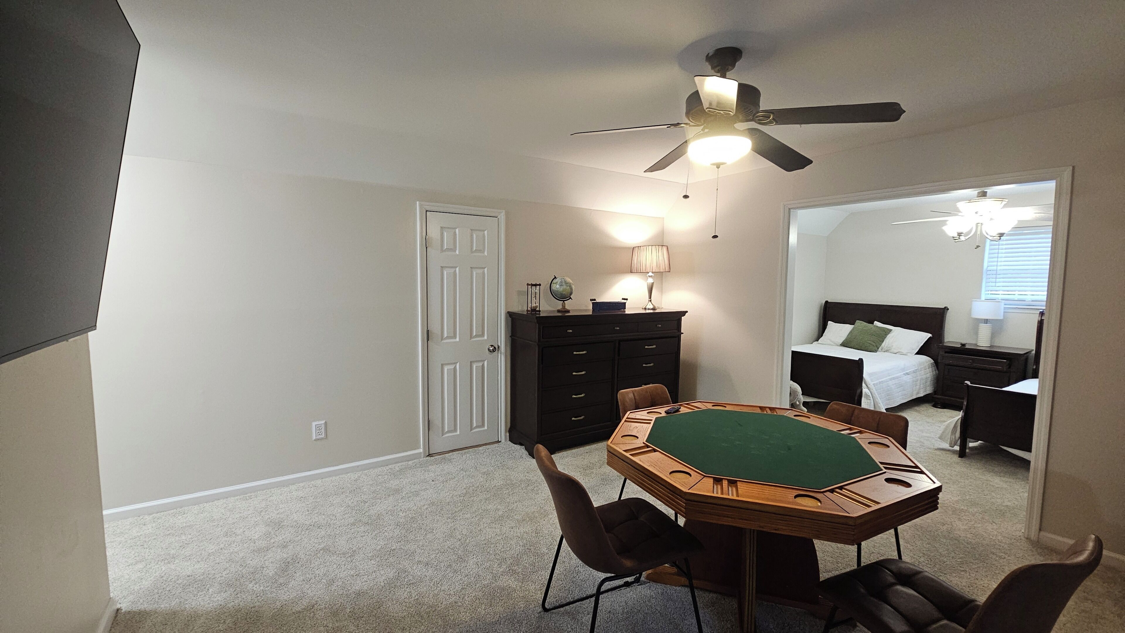 Une chambre à coucher comprenant un lit, une commode, un bureau avec un plateau recouvert de feutre vert, un ventilateur de plafond et une porte.