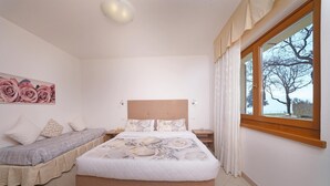 Ein Schlafzimmer mit einem großen Bett, einem kleineren Bett, einem Nachttisch, einem Fenster mit Vorhängen und einer Wand mit einem floralen Gemälde.