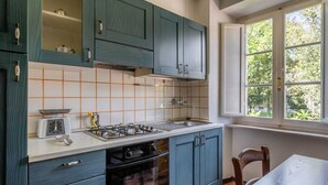 Una cocina con armarios azul verdoso, encimera blanca, estufa a gas, un lavamanos y una ventana con vistas a vegetación.