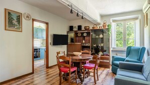 Un comedor con una mesa y sillas de madera, un sillón azul, y una cocina visible a través de un umbral abierto.