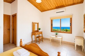 A bedroom with a wooden ceiling, a bed with a wooden headboard, a dresser with a mirror, a small table with a plant, a window with a view of the sea, and an air conditioning unit. - Villa Chrisandria - Three Bedroom Villa, Sleeps 6 (Argaka)