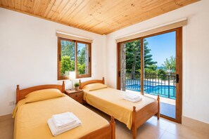 A bedroom with two beds, a nightstand, a lamp, and a view of a swimming pool. - Villa Chrisandria - Three Bedroom Villa, Sleeps 6 (Argaka)
