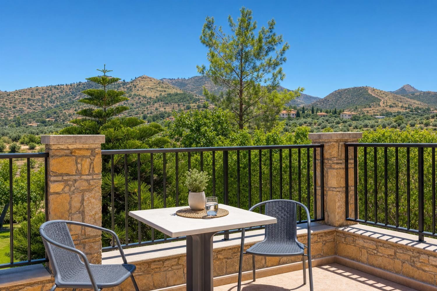 A balcony with stone pillars, two chairs, a table, and a view of a mountainous landscape.