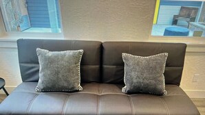 Un sofá de cuero marrón oscuro con dos almohadas grises, ubicado en una habitación con un ventanal y vistas a edificios.