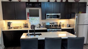 Cocina moderna con armarios oscuros, una isla central con sillas, un refrigerador de acero inoxidable y un microondas sobre la estufa.