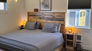 Un dormitorio con cama, mesitas de noche, ventilador de techo, ventana con persianas y un cuadro en la pared.