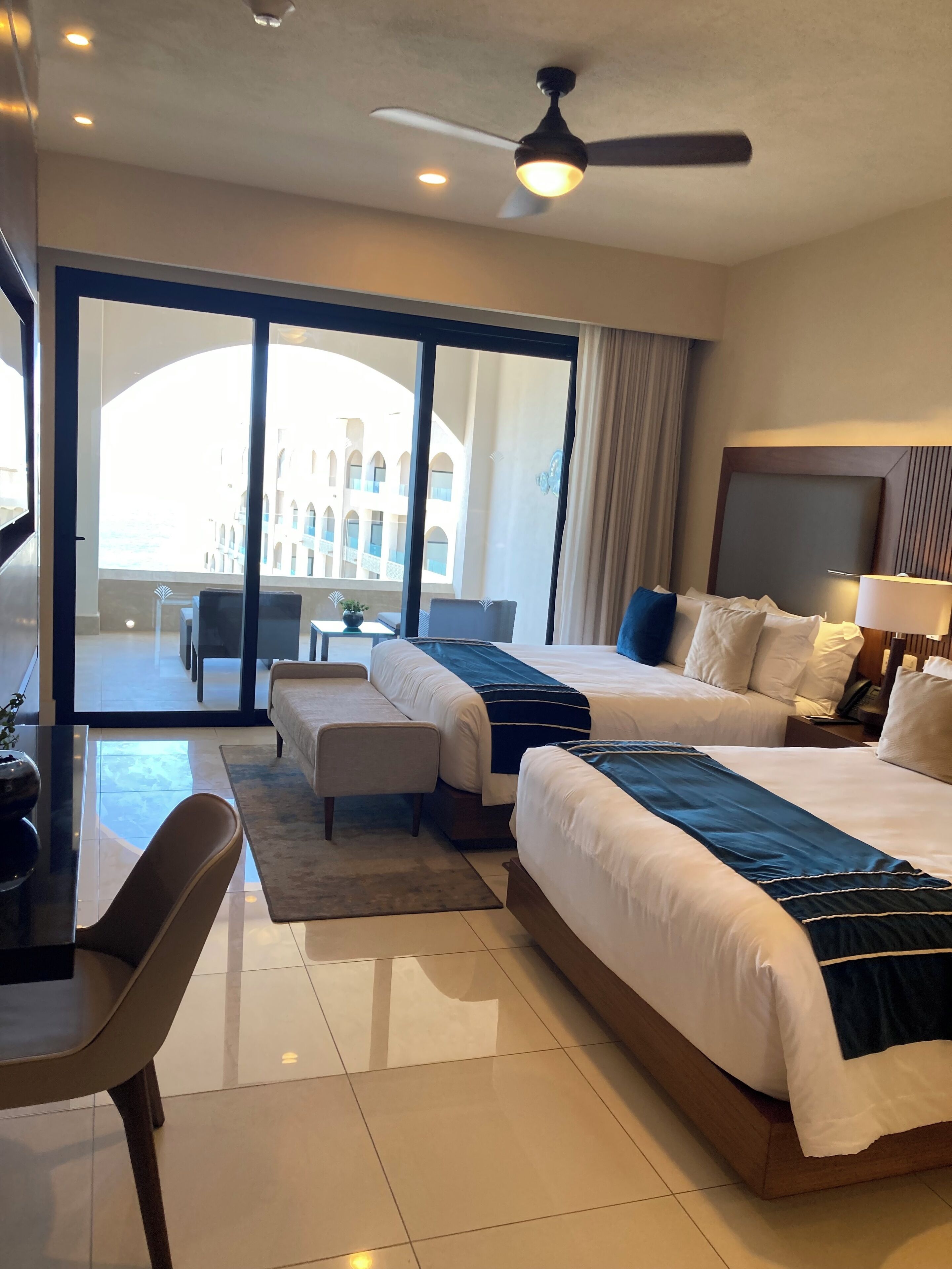 A hotel room with two beds, a desk, a chair, and a view of a building through large windows.