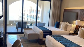 Habitación de hotel con dos camas, un escritorio, una silla y vistas a un edificio a través de amplios ventanales.