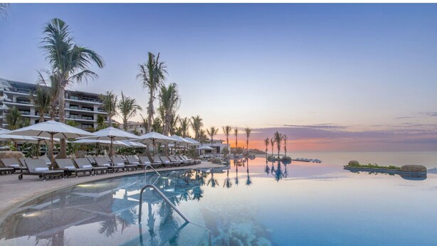 Piscina de un resort con sillas de descanso y sombrillas, rodeada de palmeras y un edificio al fondo.