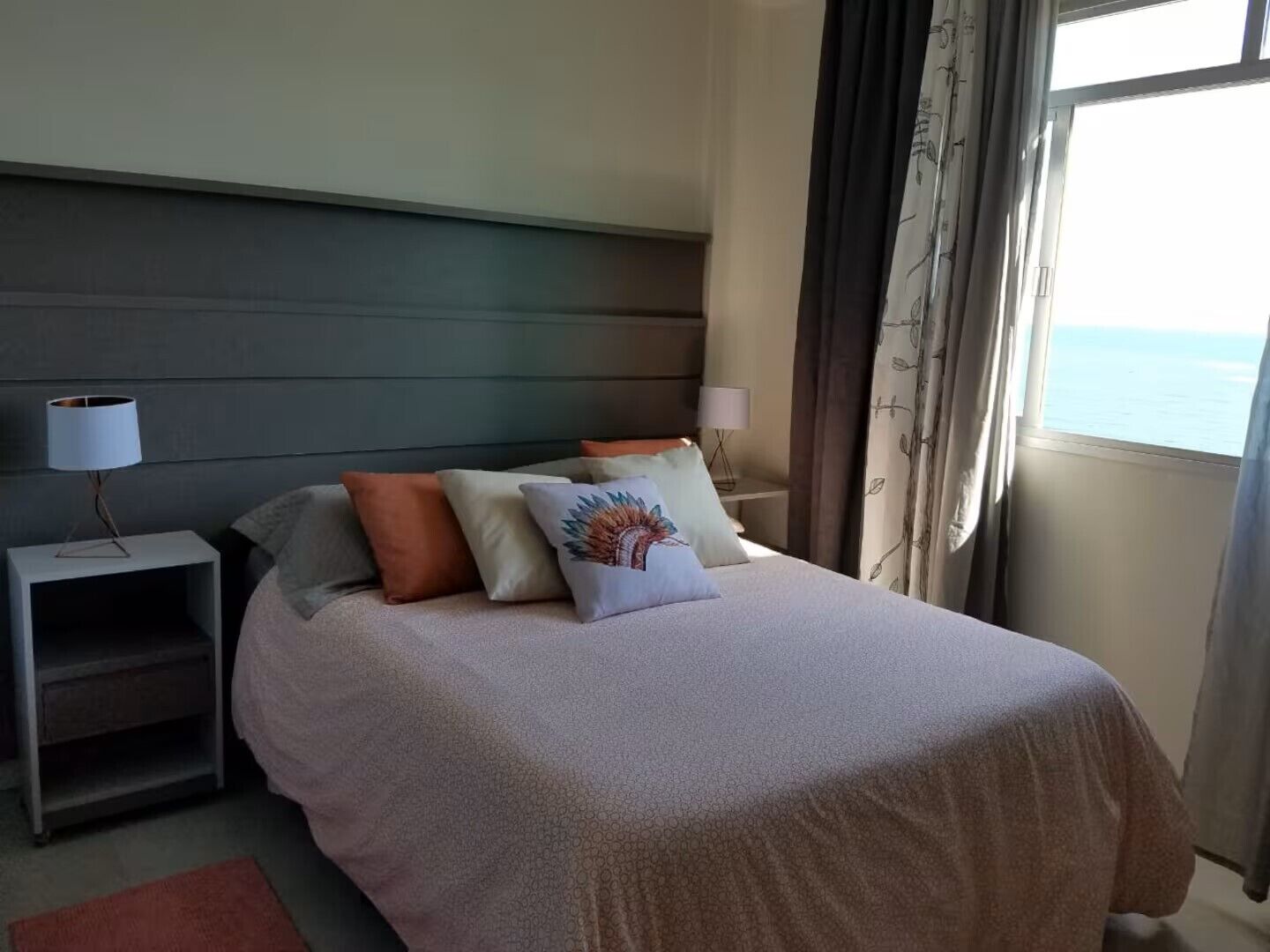 En pænt redt seng med et mønstret sengetæppe og flere puder, et natbord med en lampe, og havudsigt gennem et vindue med gardiner.