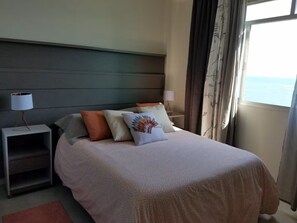 A neatly made bed with a patterned bedspread and several pillows, a bedside table with a lamp, and a view of the sea through a window with curtains. - Beautiful beachfront apartment in Balneário Camboriú. Cozy with a fantastic view. (Balneário Camboriú)