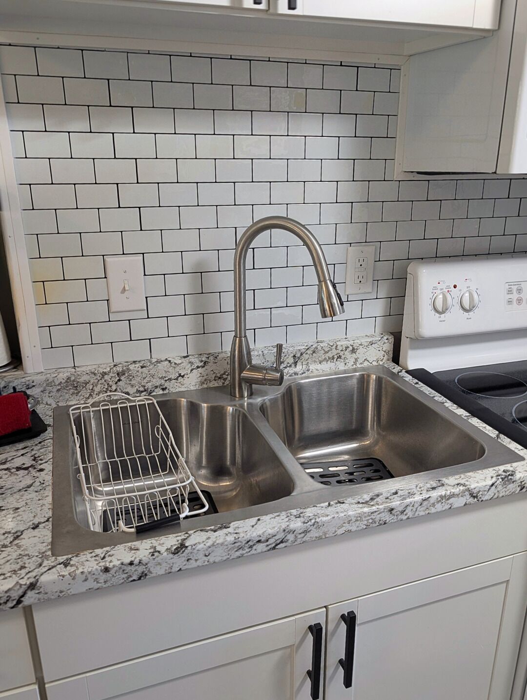 A kitchen with a double stainless steel sink, a dish rack, a white tiled backsplash, and a white stove.