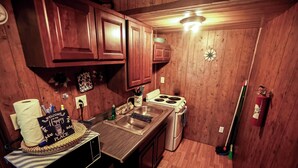 Cocina compacta con muebles de madera, un lavamanos de acero inoxidable, una estufa y una escoba apoyada en la pared.
