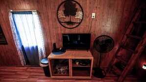 Interior de madera con un televisor sobre un pedestal, un ventilador y una estantería con objetos.