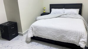 Una cama bien hecha con ropa blanca, una mesita de noche negra con lámpara, y un mueble de almacenamiento negro con cajones.