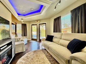 A modern living room with a beige sofa, a flat-screen TV, and a fireplace. There is a ceiling with blue lighting and a sign indicating an exit. - Haven at Hazel - the Residence.  A unique stay overlooking the Demorest Square. (Demorest)