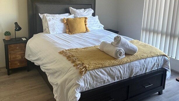A neatly made bed with white and yellow bedding, a dark headboard, and a bedside table with a lamp and a small plant. - 10 mins drive to T4 terminal, walking distance to the Swan river! Family home. (Bayswater)