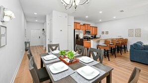 A modern kitchen with wooden cabinets, a dining area with a table set for four, and a blue sofa in the living room. - Bright & Breezy Disney Escape | Pool | BBQ (Kissimmee)