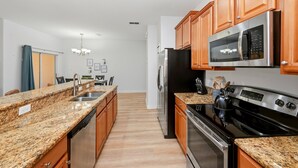 A kitchen with granite countertops, wooden cabinets, stainless steel appliances, and a dining area in the background. - Bright & Breezy Disney Escape | Pool | BBQ (Kissimmee)